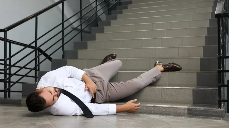 Man in business attire lying on floor at bottom of staircase, appearing injured or unconscious.