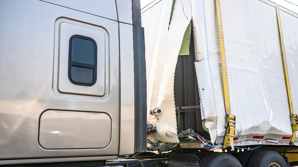 Close-up of a silver truck cab and a damaged white trailer with torn side panels and yellow straps.