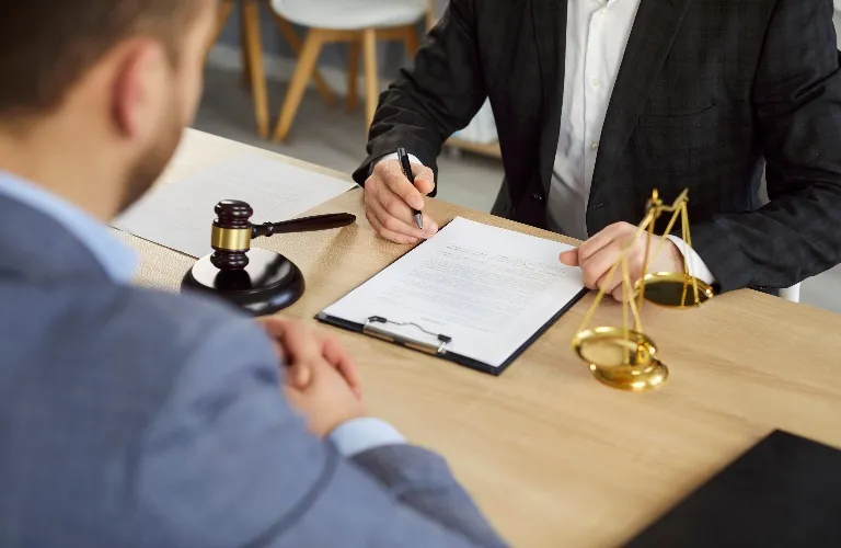 Two men in formal attire at a desk with legal documents, a gavel, and scales of justice.