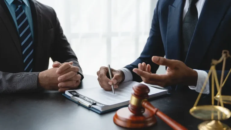 Two men in suits discussing legal documents with a gavel and scales of justice on the table.