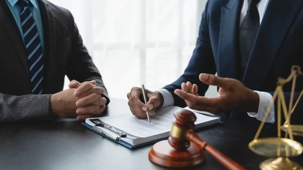 Two men in suits discussing legal documents with a gavel and scales of justice on the table.