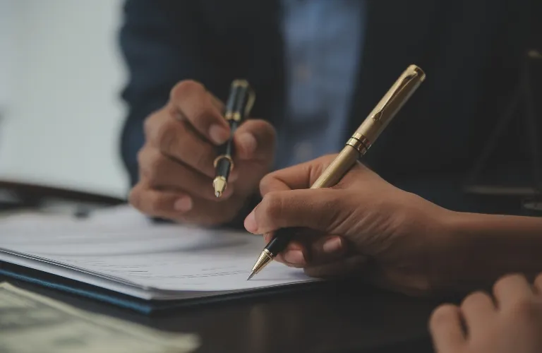 Close-up of two individuals holding pens and reviewing documents on a dark surface.