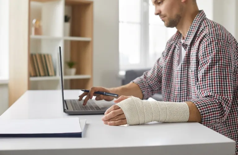 Man with a bandaged hand working on a laptop at a white desk in an office.