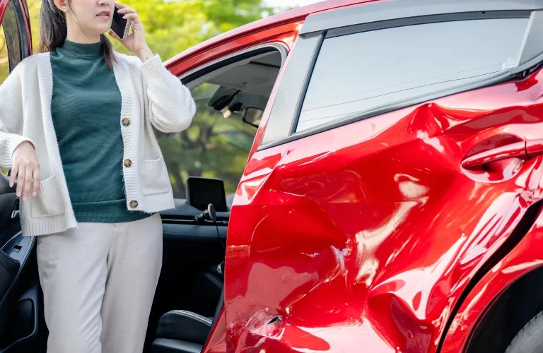 Woman in white sweater on phone next to red car with severe side door damage.