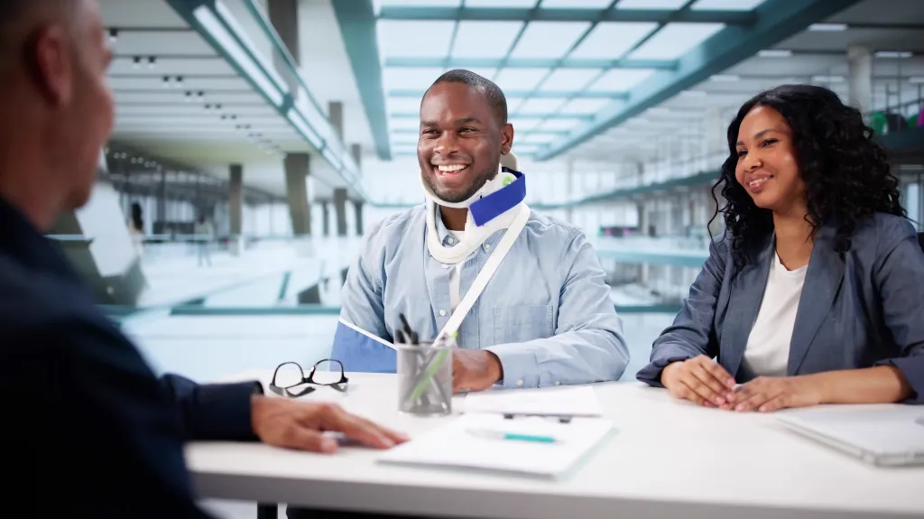 Three professionals in a modern office, one man with a neck brace and arm sling, engaged in a meeting.