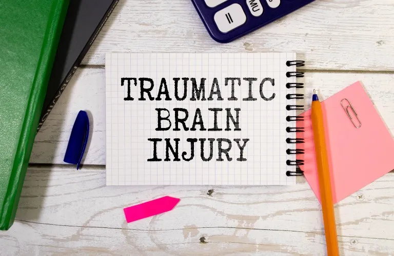 Spiral notebook with "TRAUMATIC BRAIN INJURY" text, pen, paperclip, calculator, and green book on white wooden surface.