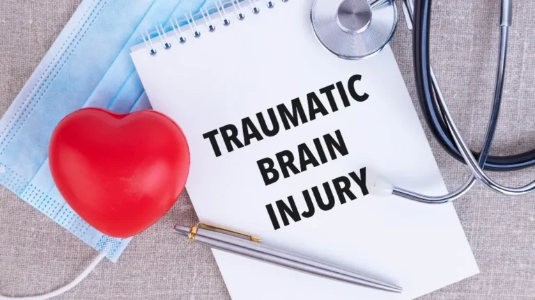 Notepad with "Traumatic Brain Injury" text, red heart model, face mask, stethoscope, and pen on fabric surface.