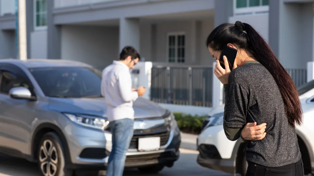 Two individuals using mobile phones near two parked cars in a residential area.