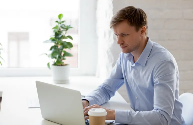 Professional man in light blue shirt working on laptop at white desk with coffee cup and green plant.