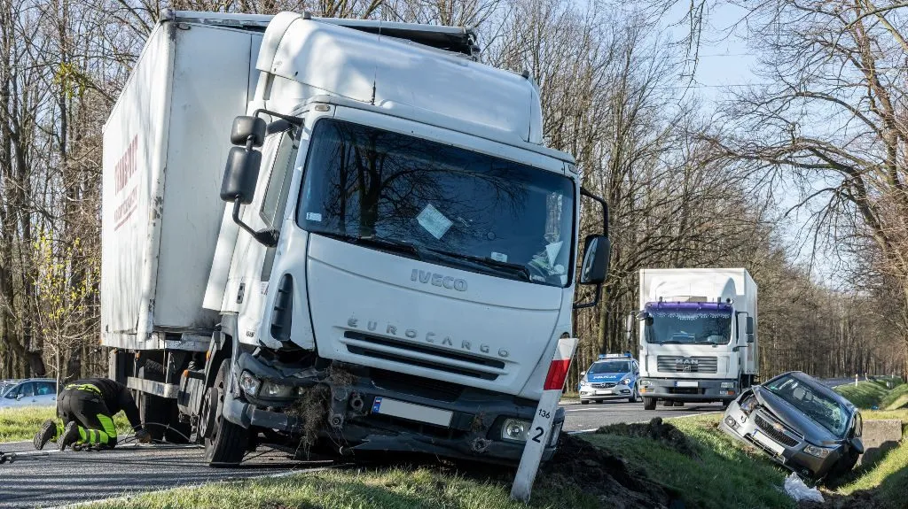 White Iveco Eurocargo truck partially off road with front damage, black car in ditch, emergency responder nearby.