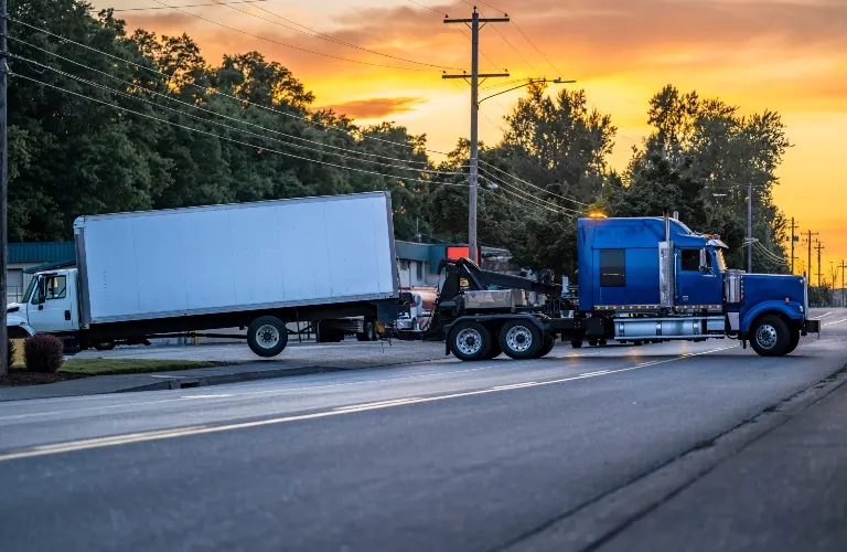 Blue semi-truck towing a white box truck on a suburban road at sunset.