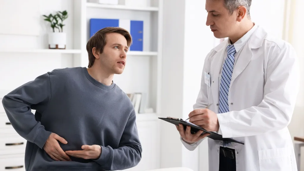 Patient in gray sweatshirt clutching abdomen while consulting with male doctor in white coat.