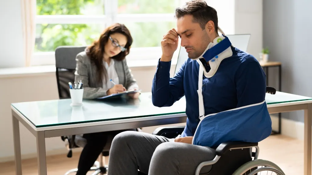 Man with neck brace and arm sling in wheelchair consulting female professional at office desk.