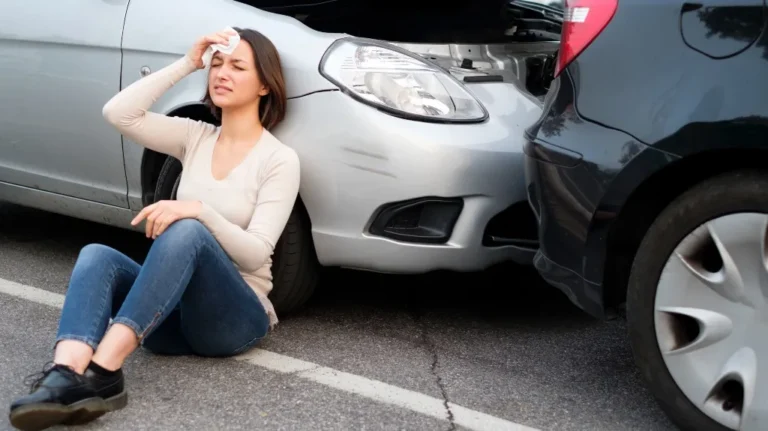 Woman in beige sweater and jeans sitting on pavement, holding head near two cars after collision.