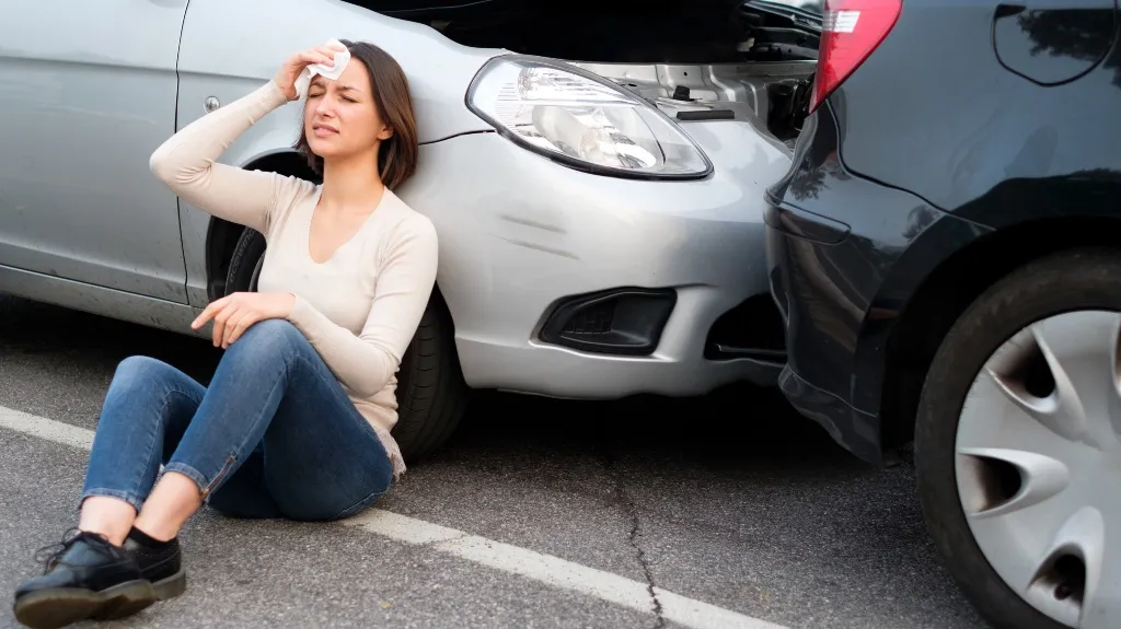 Woman in beige sweater and jeans sitting on pavement, holding head near two cars after collision.