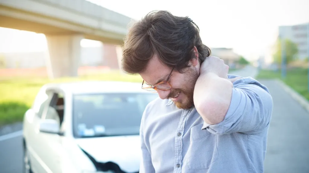 Man in light blue shirt and glasses holding neck in pain beside white damaged car.